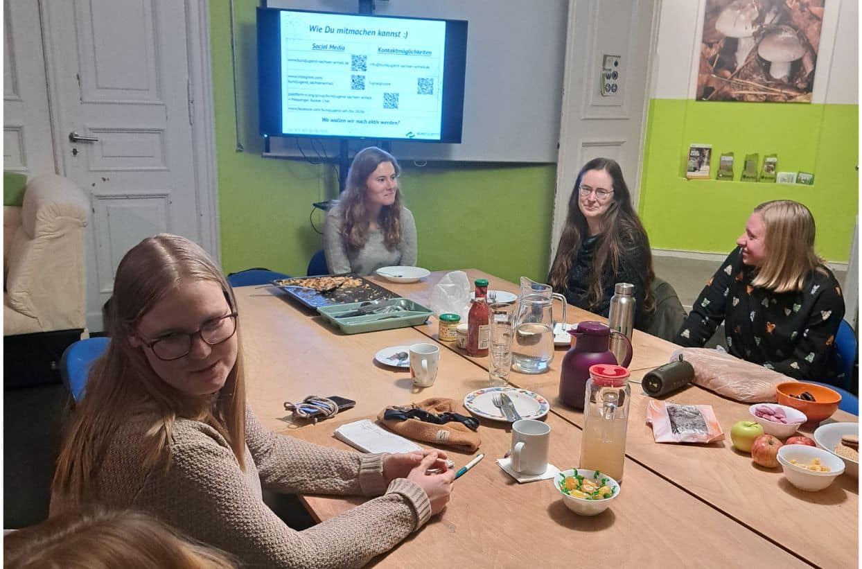 Personen im Besprechungsraum mit Essen und Bildschirm.